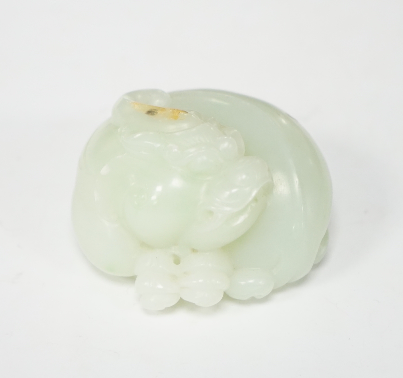 A Chinese celadon jade carving of a lion-dog, in recumbent pose, its crest with a russet inclusion, 5.3cm